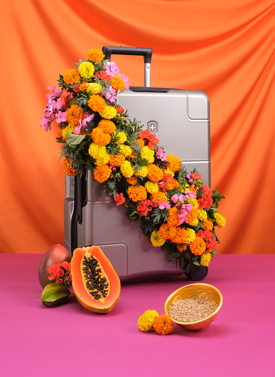 Silver suitcase decorated with vibrant orange, yellow, and pink flowers beside tropical fruits and a bowl on a pink surface.
