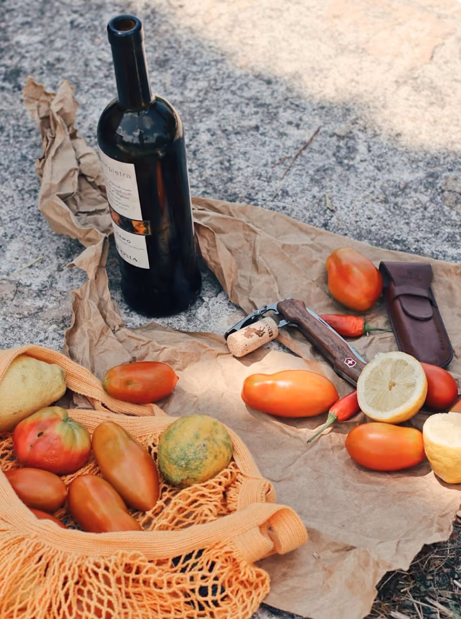 Wine bottle, tomatoes, lemons, chili peppers, and corkscrew on crumpled brown paper outdoors.