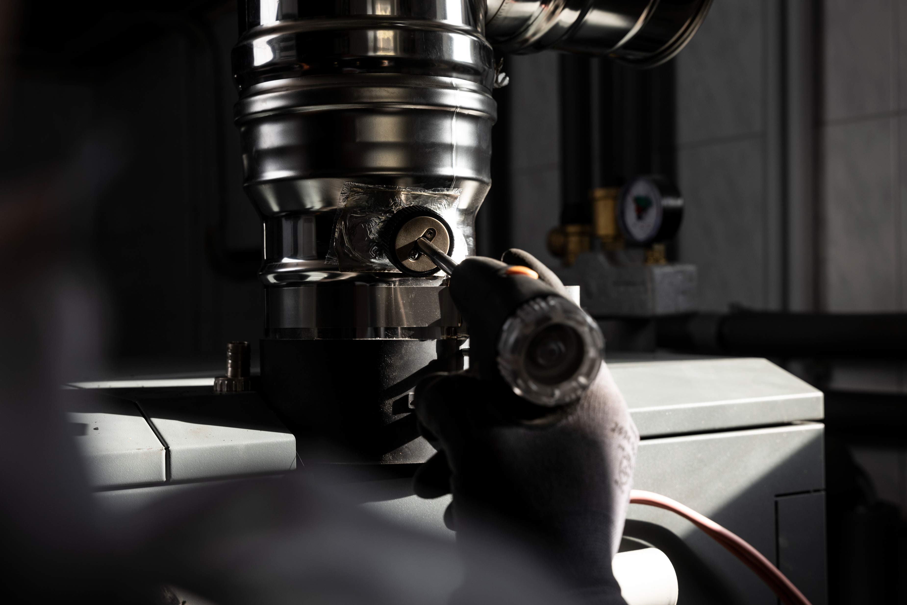 Person wearing gloves using a screwdriver to adjust a metal pipe connection on a heating system.