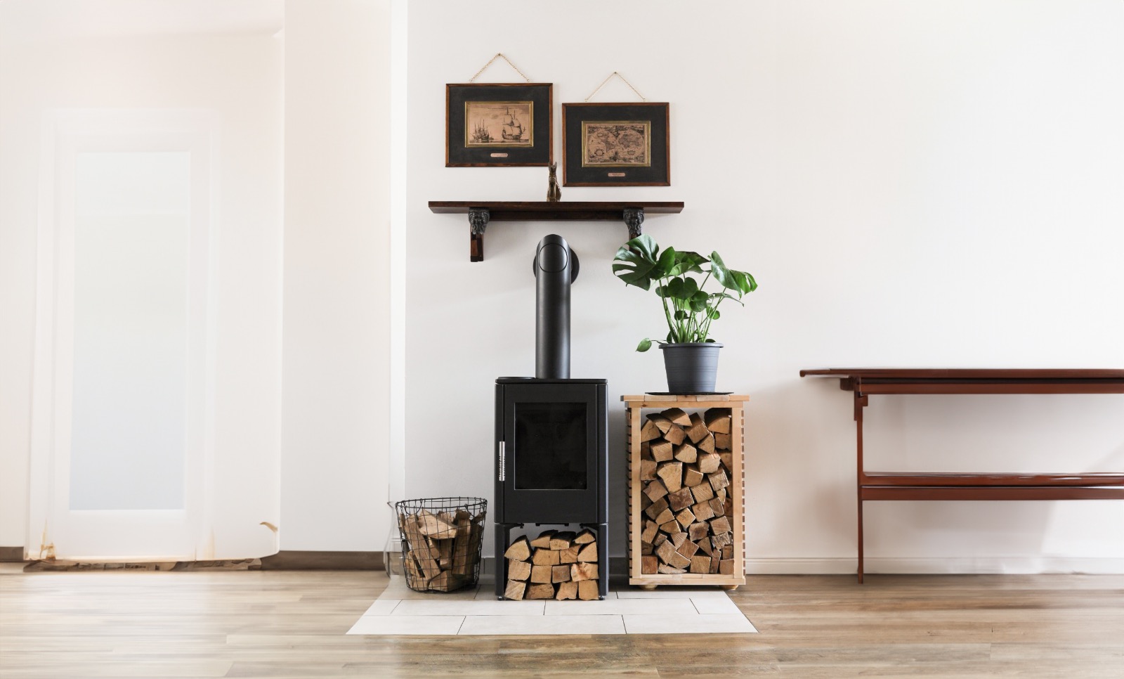 Modern black wood-burning stove with stacked firewood in baskets on each side, a green potted plant on a wooden stand, and two framed vintage artworks on a wall-mounted shelf above.