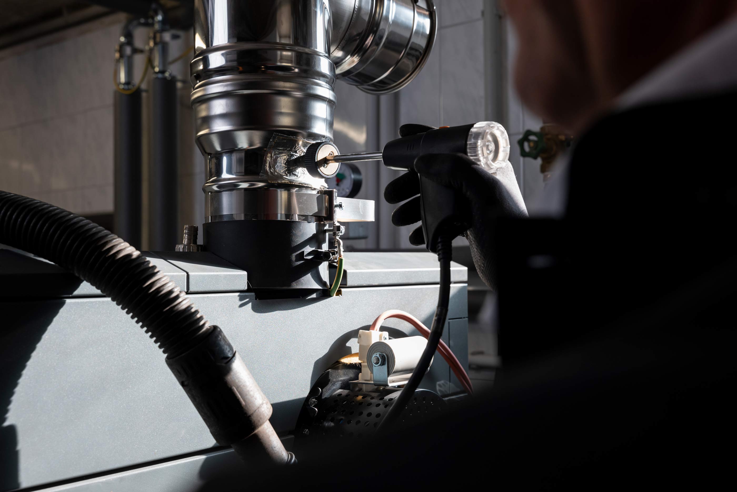 Technician wearing black gloves using a diagnostic tool on industrial piping and machinery in a workshop.