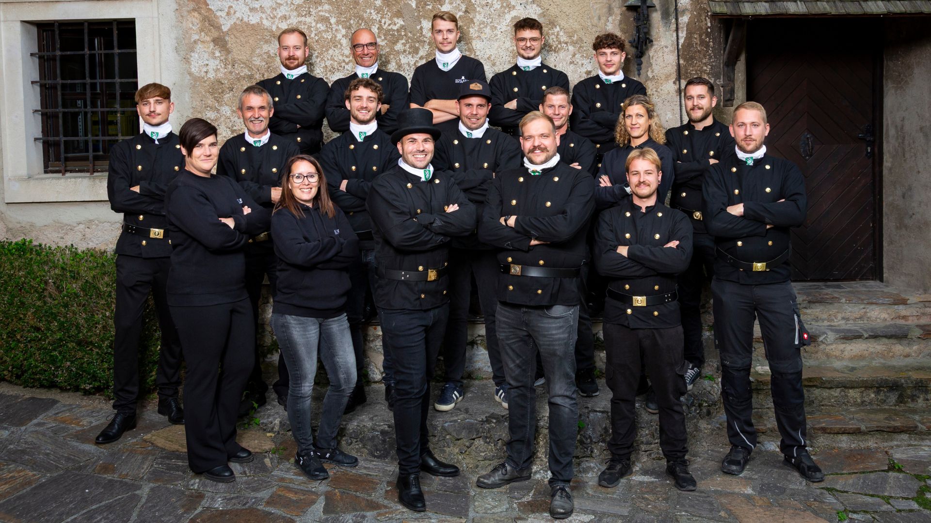 Group of 19 people standing and posing outdoors in front of an old textured wall, most wearing black uniforms with golden buttons and some in casual black clothing.