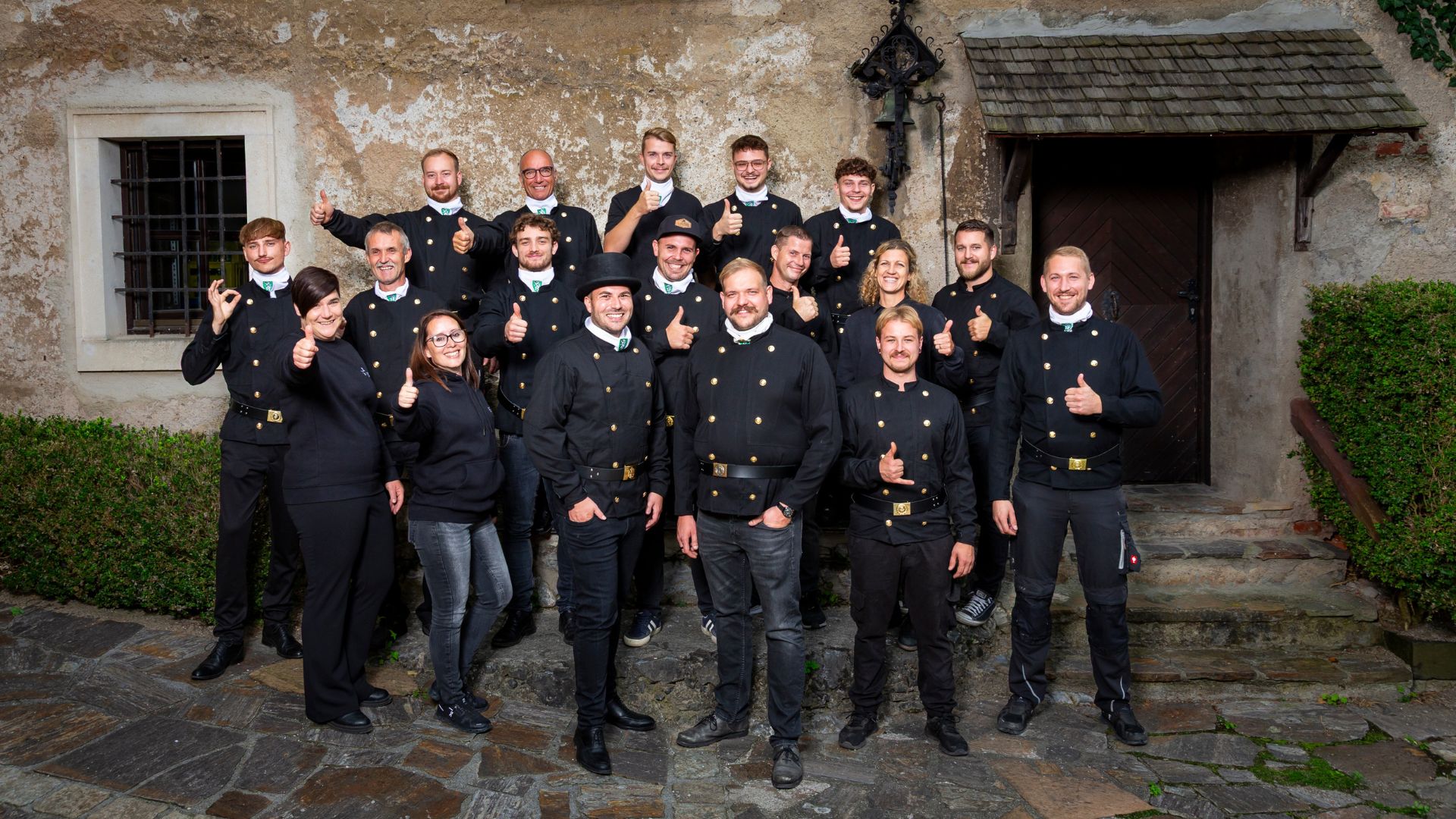 Group of people wearing black uniforms and casual clothes posing outdoors in front of a rustic stone wall, many showing thumbs up.