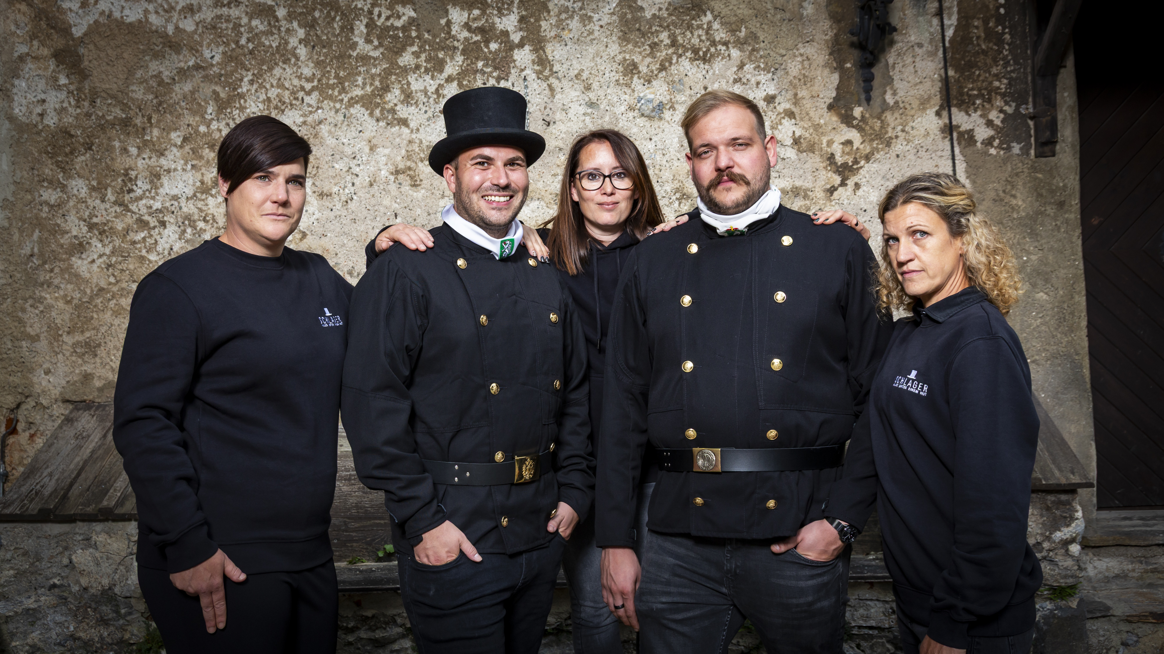Group of five adults standing against a textured wall, with two men in black uniforms with gold buttons and a top hat, and three women in black casual clothing.