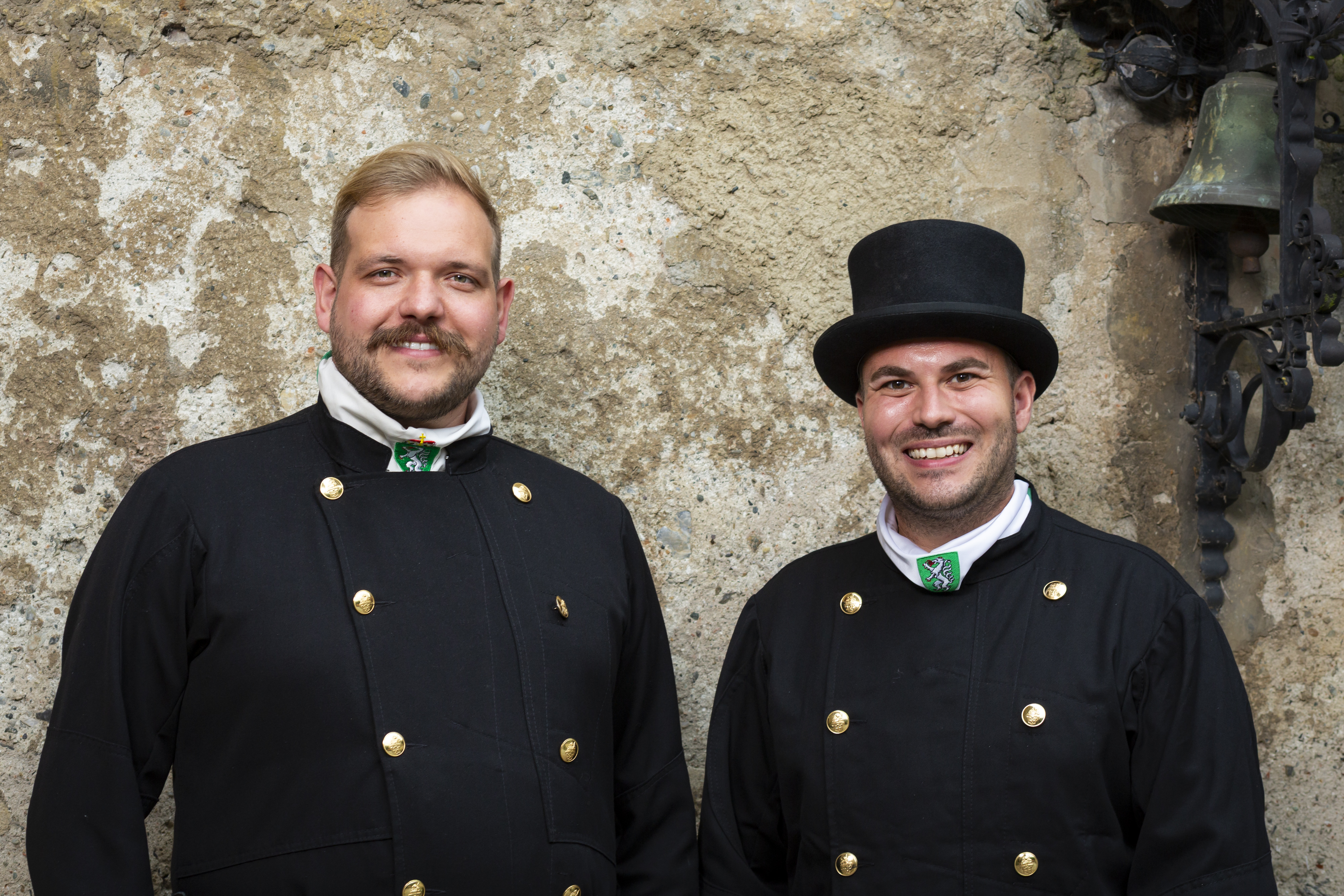Two smiling men wearing black traditional uniforms with gold buttons and green emblem collars, one wearing a black top hat, standing against a textured stone wall.