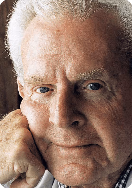 Close-up of an elderly man with white hair and blue eyes resting his face on his hand.