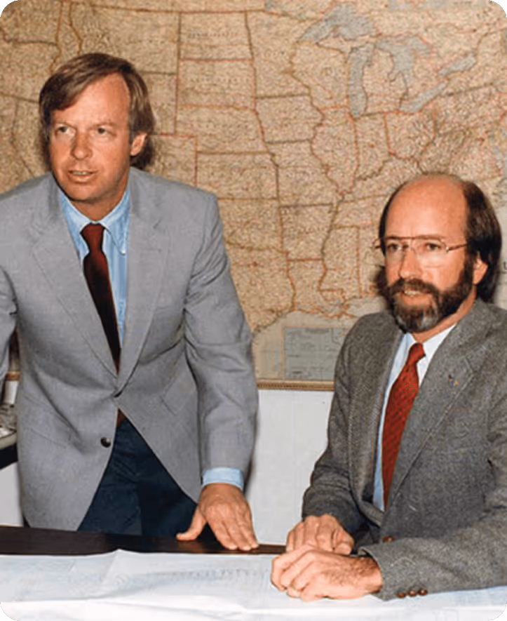 Two men in suits with red ties standing and sitting in front of a map of the United States.