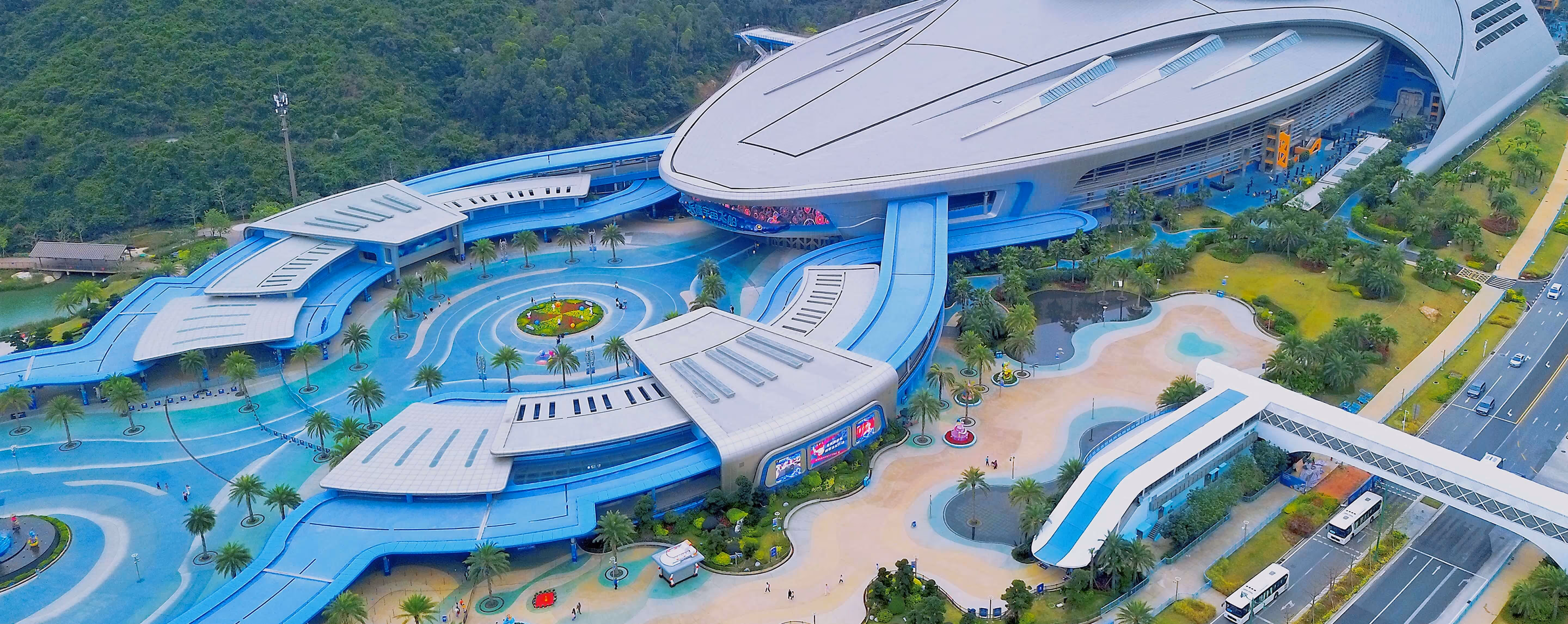 Aerial view of a large, futuristic building complex with blue and gray curved structures, surrounded by palm trees, walkways, and greenery.