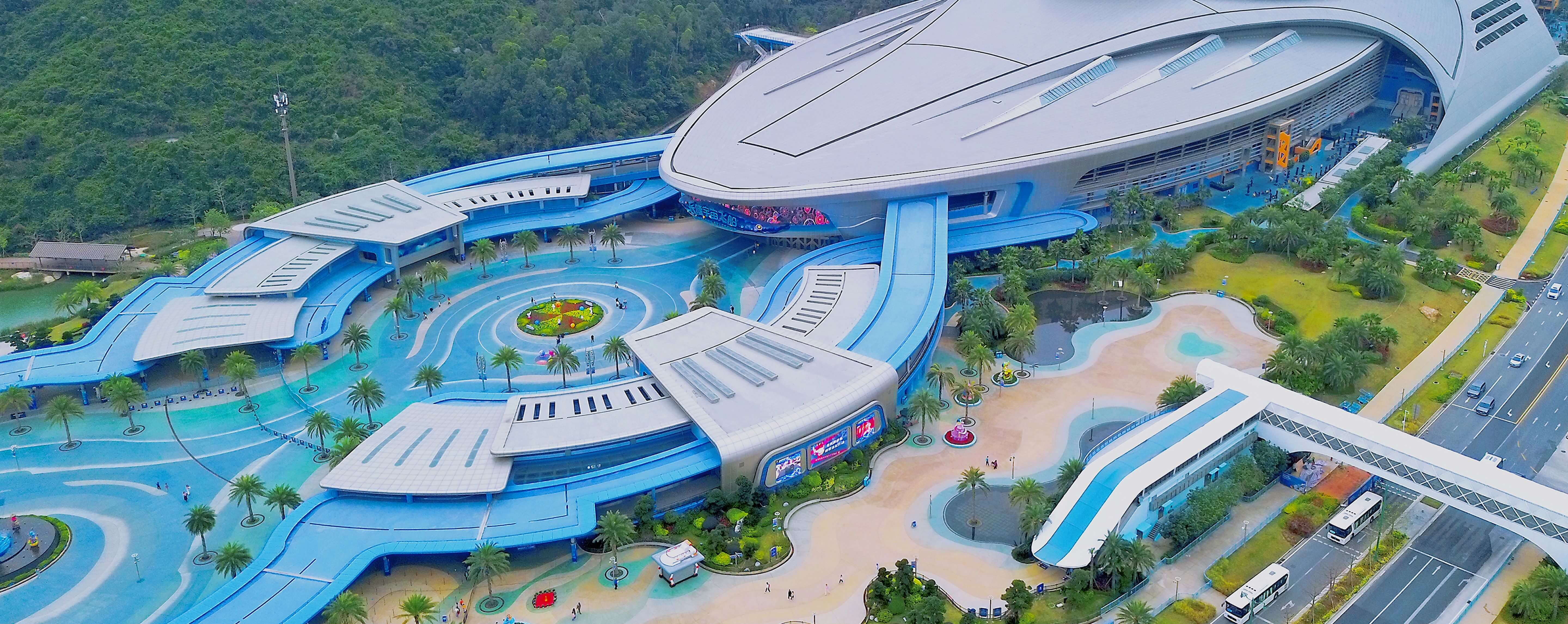 Aerial view of a large, futuristic building complex with blue and gray curved structures, surrounded by palm trees, walkways, and greenery.