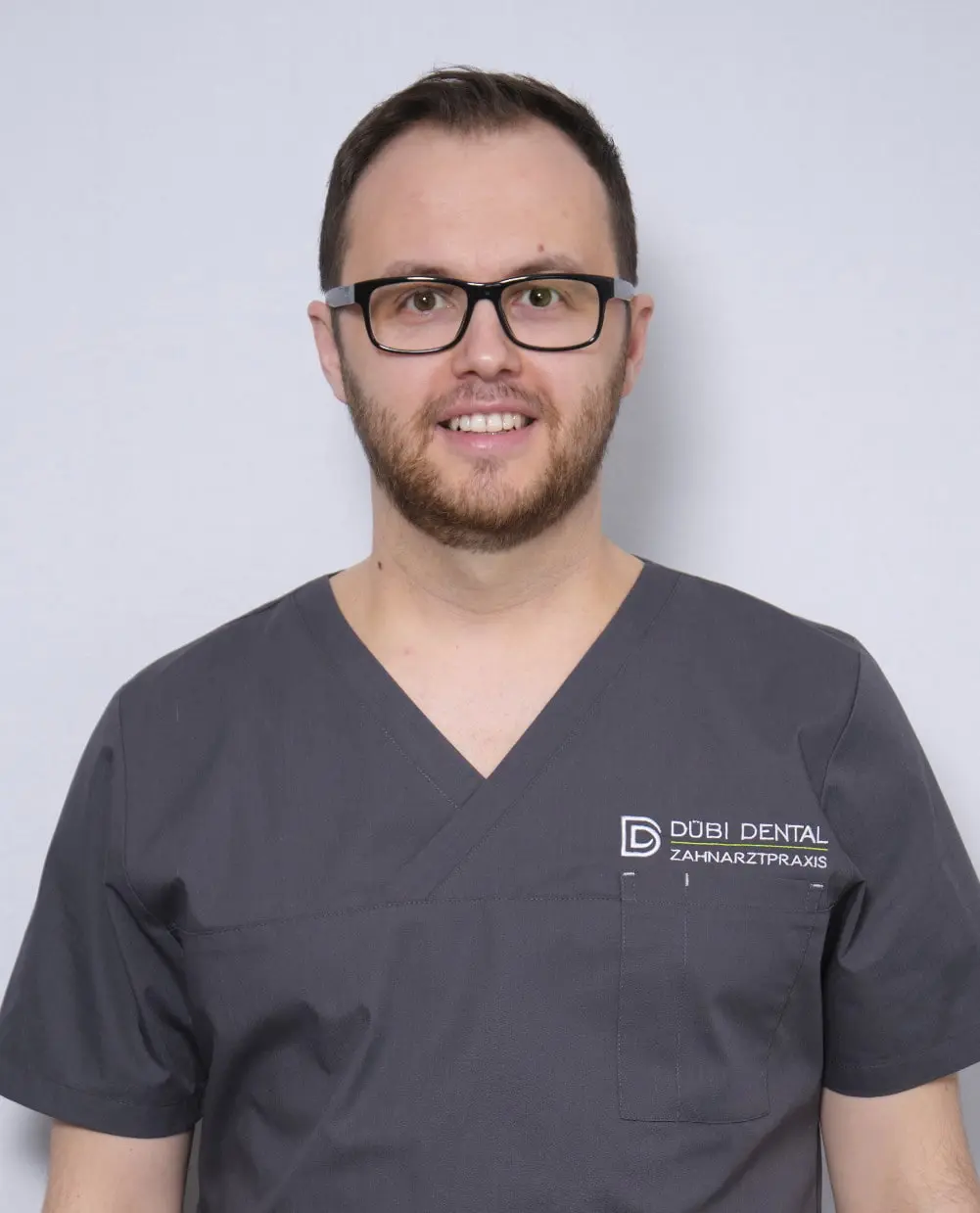 Mann mit Brille und Bart trägt ein dunkelgraues medizinisches Oberteil mit der Aufschrift Dübi Dental Zahnarztpraxis.