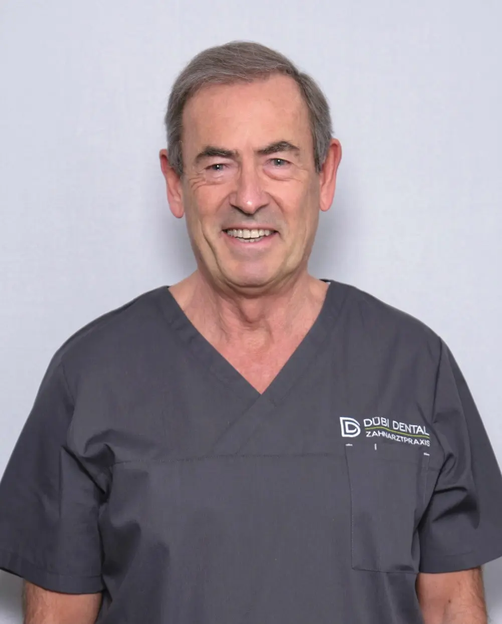 Lächelnder älterer Mann in grauer Zahnarzt-Uniform mit Logo von Dübi Dental auf hellem Hintergrund.