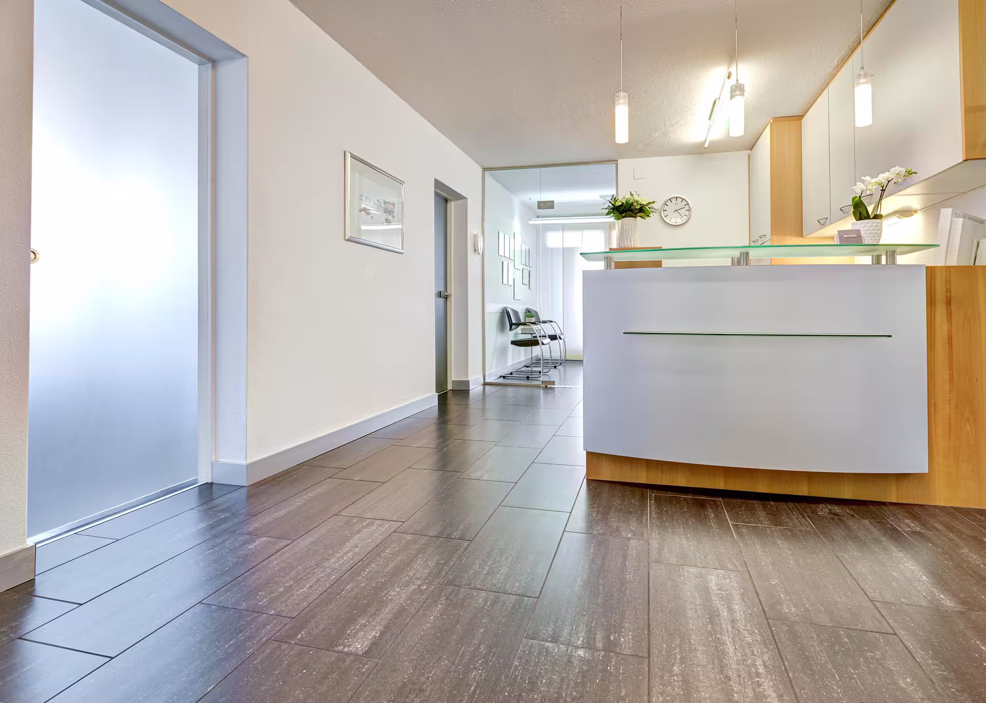 Heller, moderner Empfangsbereich mit weißem Tresen, Holzakzenten, Glaswand und Sitzgelegenheiten hinter dem Empfang.