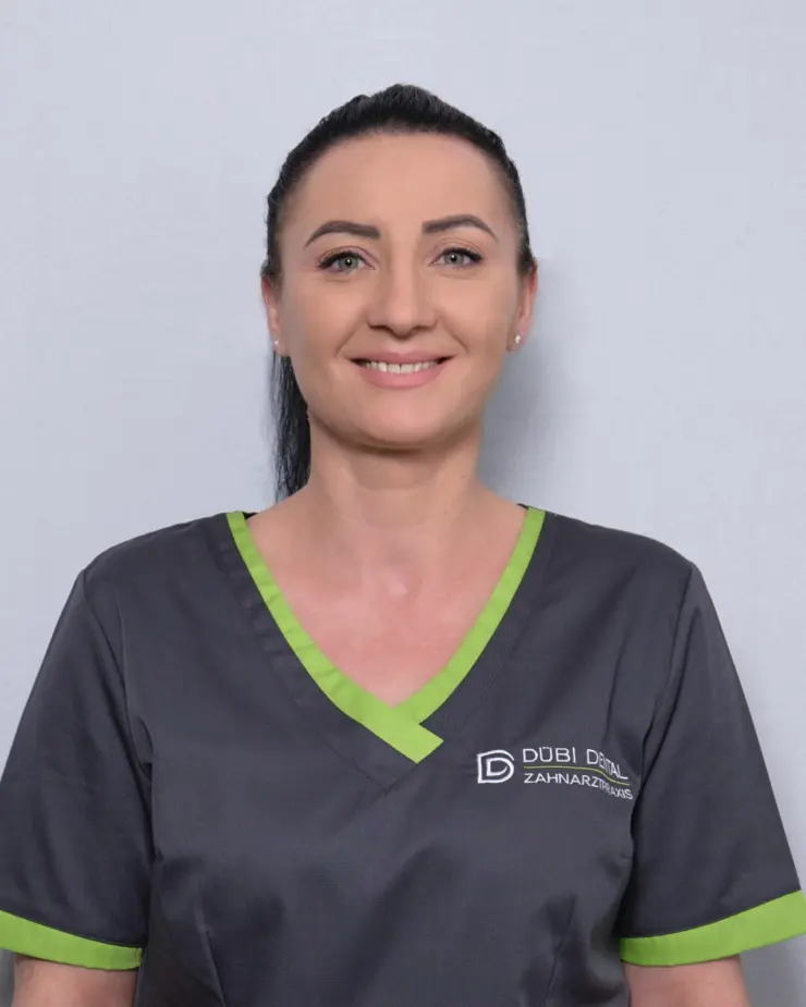 Lächelnde Frau mit zusammengebundenem schwarzen Haar trägt eine graue Arbeitskleidung mit grünen Akzenten und dem Logo von Dübi Dental Zahnarztpraxis.