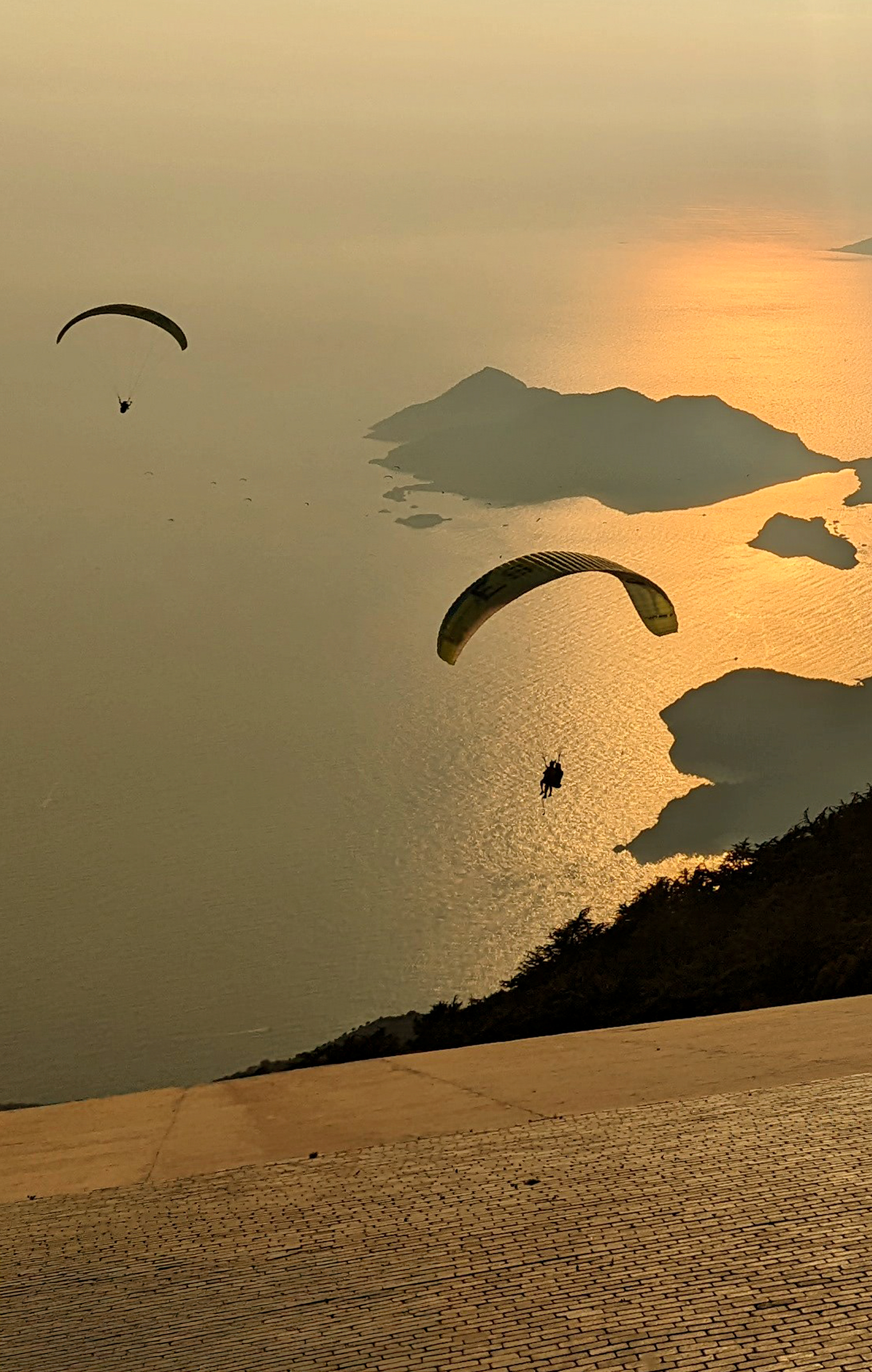 Two paragliders flying over a coastline at sunset with golden light reflecting on the water.