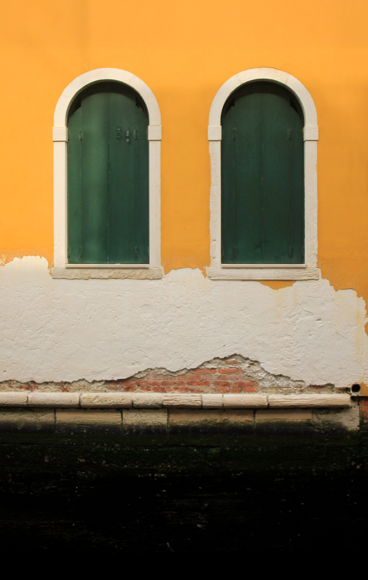 Two green arched window shutters on a weathered yellow and white wall above dark water.