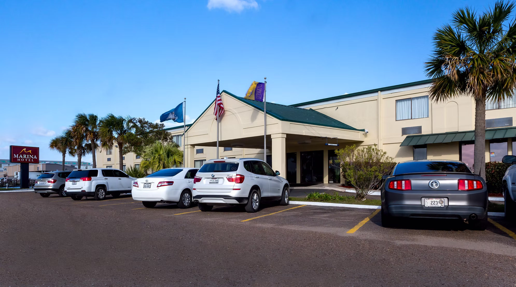 Exterior view of Marina Motel with parked cars, palm trees, and three flags in front against a clear blue sky.