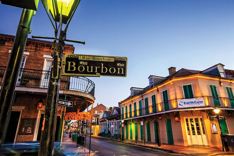 Bourbon Street sign hanging from a lamppost at dusk, with colorful buildings and street lights lining the street.