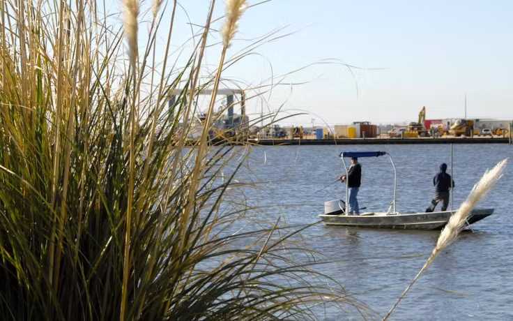 Two people fishing from a small boat on a calm river with tall grasses in the foreground and industrial equipment visible on the opposite shore.