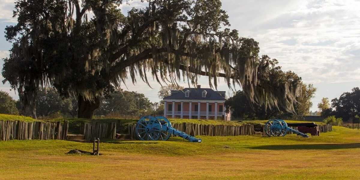 Historic battlefield with two blue cannons, wooden fortifications, a large tree draped in Spanish moss, and a pink antebellum mansion in the background.
