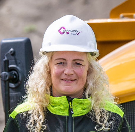 Kvinne i byggearbeidsklær med hjelm foran anleggsmaskin.