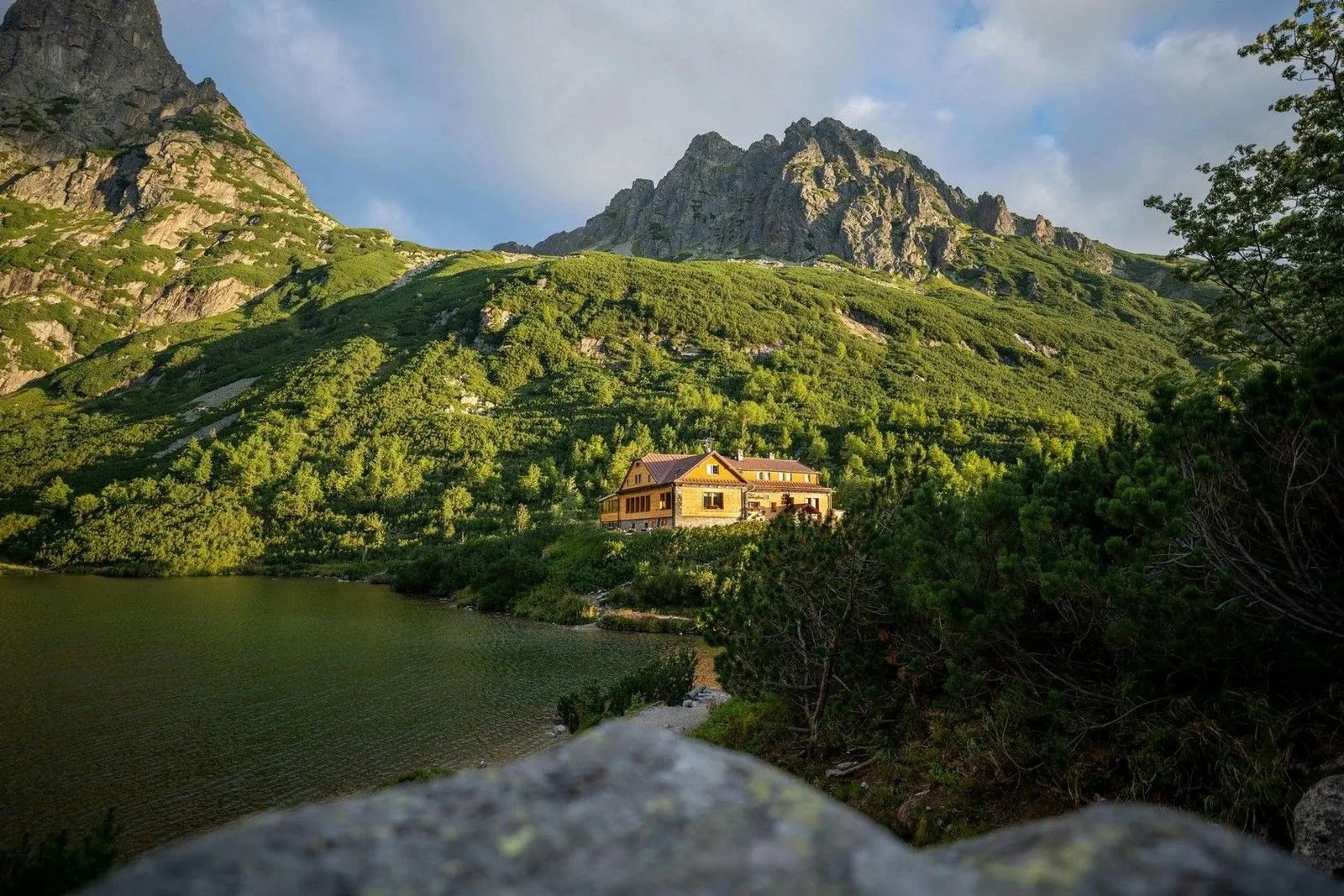 A picturesque mountain lodge by a serene lake, surrounded by lush greenery and towering rocky peaks under a cloudy sky.