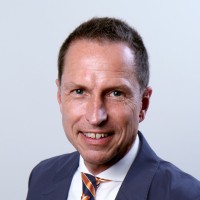 Portrait of a man in a navy suit, white shirt, and striped tie smiling against a light gray background.