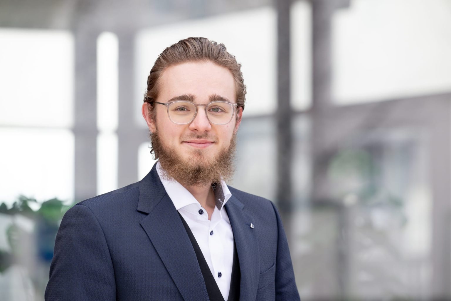 Young man with glasses and a beard wearing a navy suit and white shirt, smiling in a bright indoor setting.