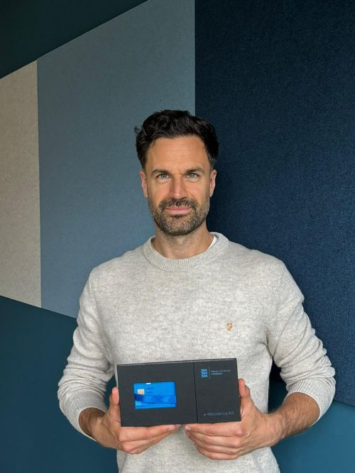 Man with dark hair and beard wearing a light gray sweater holding an Estonia e-Residency kit with a blue card inside.