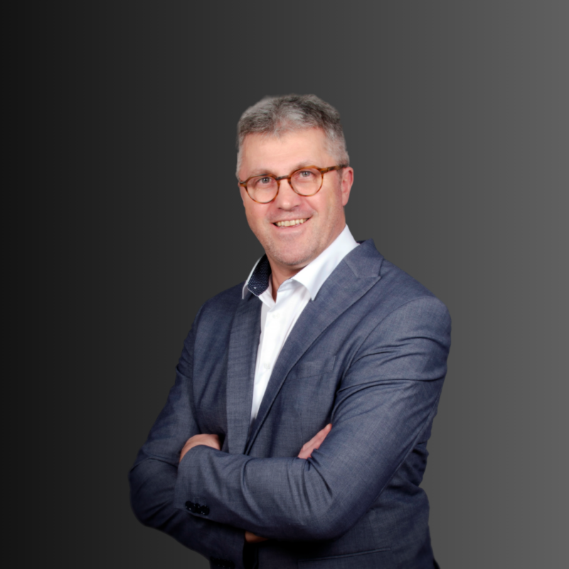 Smiling man with glasses and gray hair in a light blue shirt standing outdoors with arms crossed near a stone wall.
