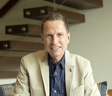 Portrait of a man in a navy suit, white shirt, and striped tie smiling against a light gray background.