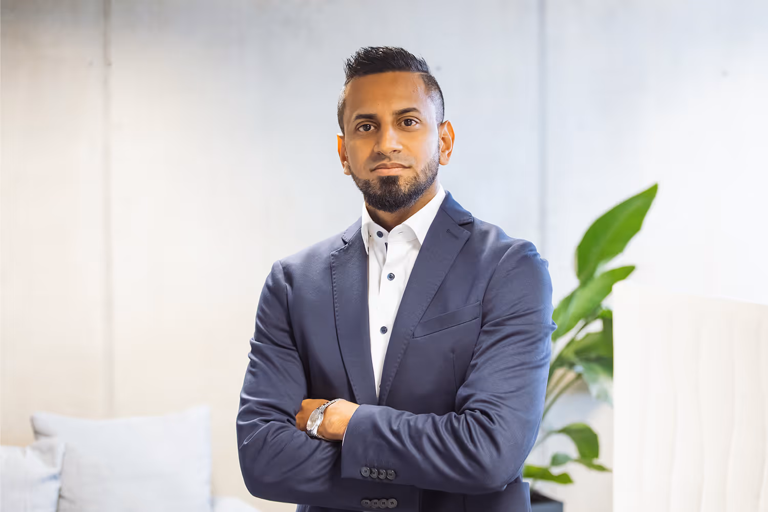 Porträt eines Mannes mit dunklem Haar und Bart, der in einem weißen Hemd und einem dunkelblauen Anzug mit verschränkten Armen vor einem modernen, hellen Hintergrund steht.