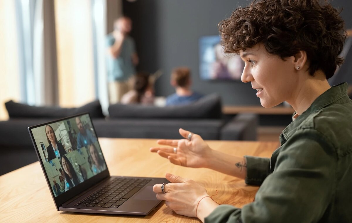 Eine Frau mit lockigen Haaren gestikuliert während einer Videokonferenz an ihrem Laptop in einem hellen Wohn- oder Arbeitsbereich.