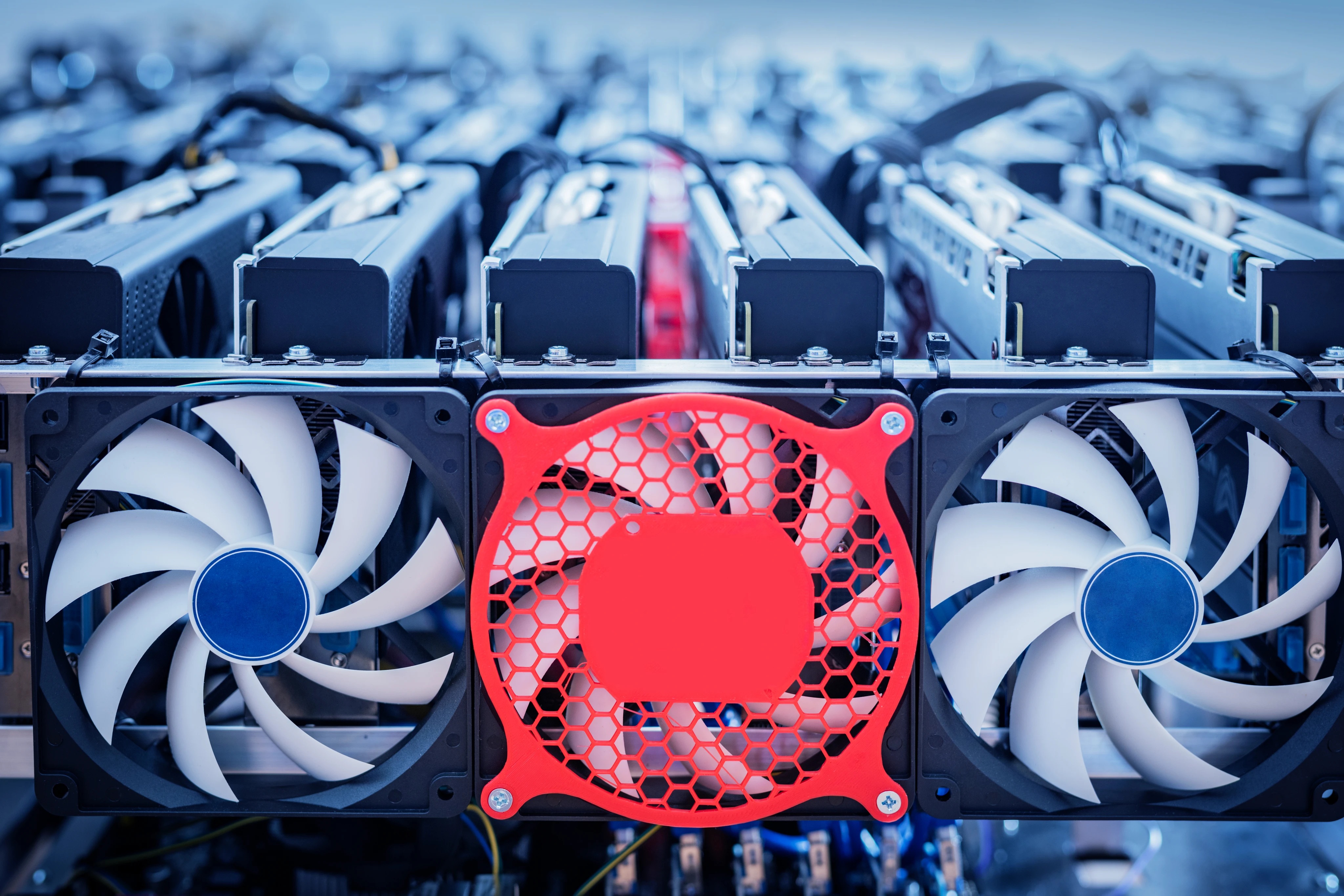 Eine Nahaufnahme zeigt eine Reihe von Lüftern an einem Krypto-Mining-Rig. Im scharfen Vordergrund sind drei große Kühllüfter zu sehen, wobei der mittlere ein auffälliges rotes Gitter hat, flankiert von zwei weißen Lüftern. Im unscharfen Hintergrund sind mehrere Grafikkarten (GPUs) in einem Rack für das Mining von Kryptowährungen montiert.