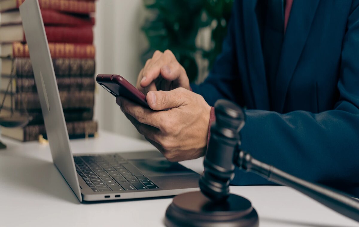 Eine Nahaufnahme aus einem juristischen Büro: Eine Person im Anzug bedient ein Smartphone über einem geöffneten Laptop. Im scharfen Vordergrund liegt ein Richterhammer, während im unscharfen Hintergrund gestapelte Rechtsbücher zu sehen sind.