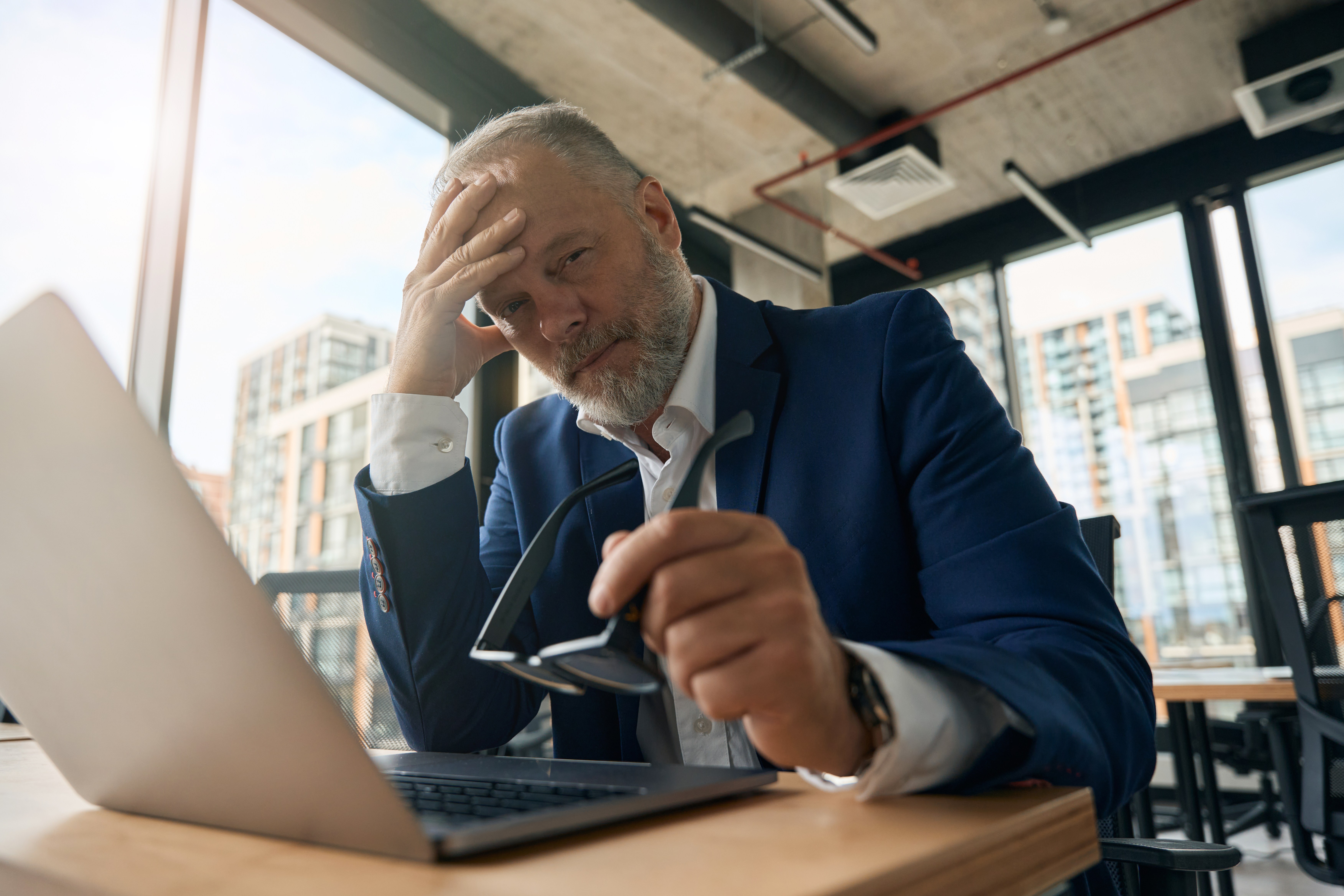Ein junger, lächelnder Mann mit lockigen Haaren und Brille sitzt an seinem Schreibtisch, hinter dem sich ein Regal mit Büchern und Dekoration befindet. Er trägt ein hellblaues Hemd über einem weißen T-Shirt und blickt direkt in die Kamera, während ein Laptop vor ihm steht.