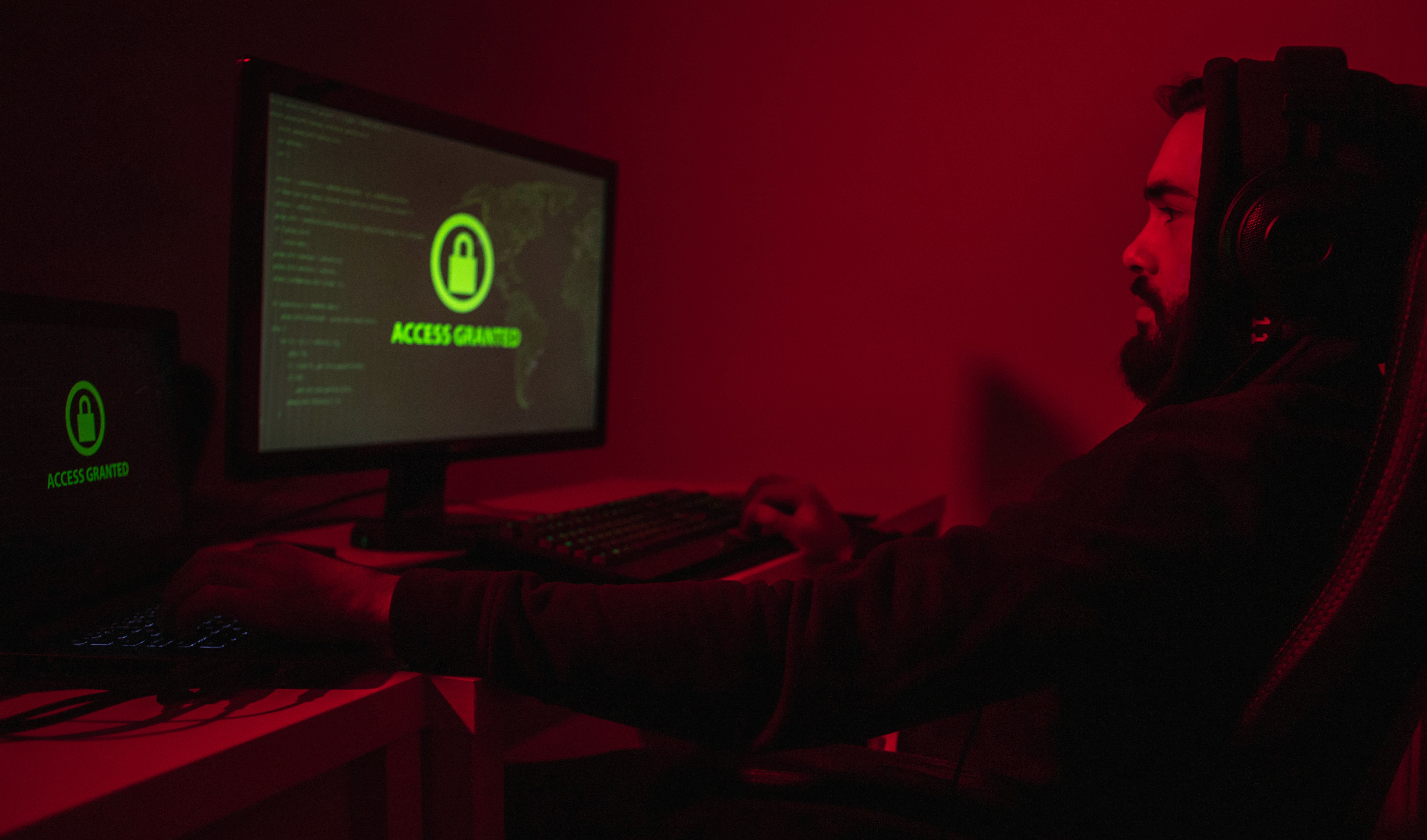Ein Mann sitzt in einem von rotem Licht beleuchteten Raum vor einem Computer. Auf dem Bildschirm ist die Meldung "ACCESS GRANTED" (Zugriff gewährt) mit einem grünen Schloss-Symbol zu sehen, was einen erfolgreichen Cyber-Einbruch signalisiert.