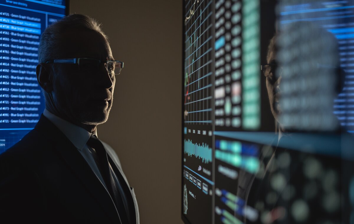 Profilansicht eines Geschäftsmannes mit Brille und Anzug, der konzentriert auf einen großen digitalen Monitor mit blauen Datenvisualisierungen und Systemmetriken blickt.