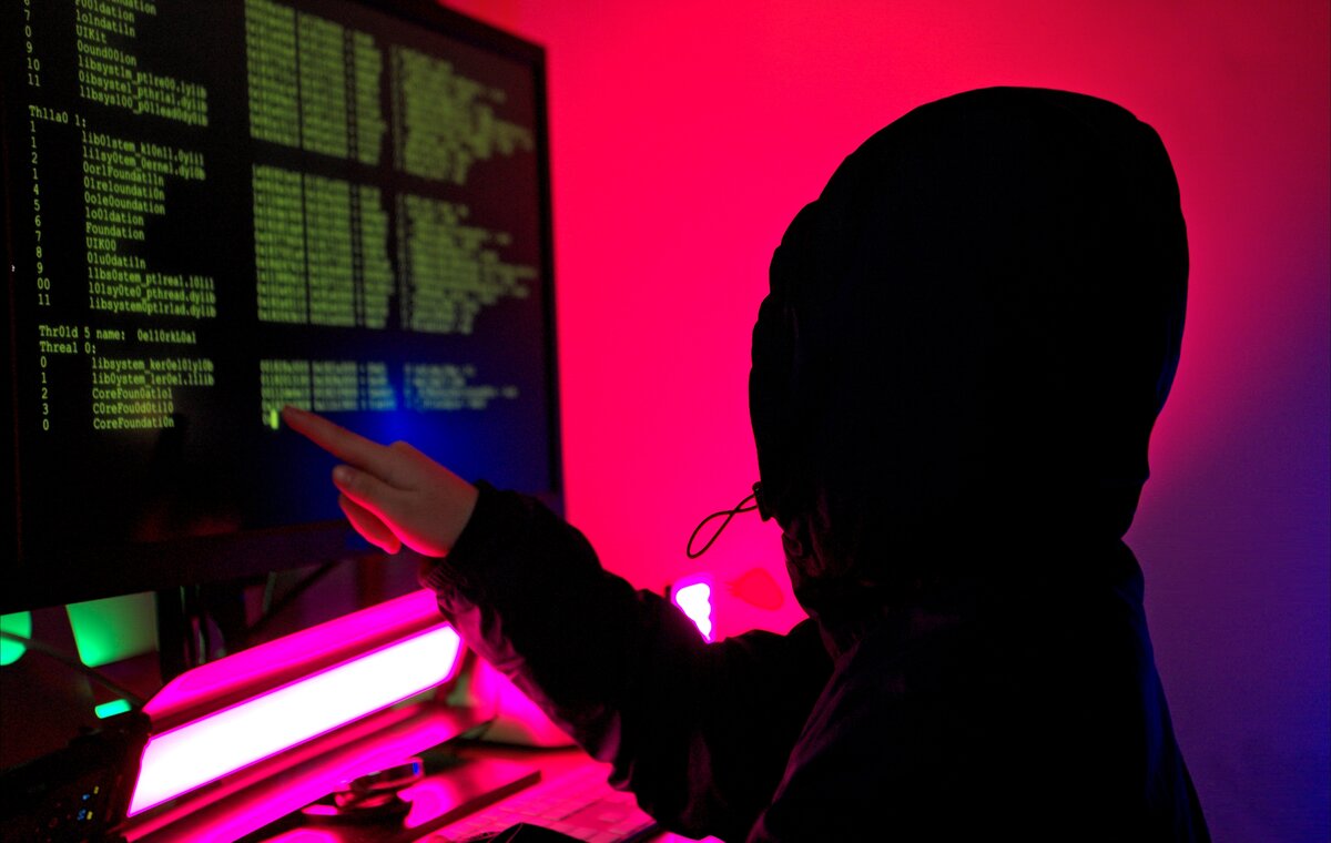 Silhouette einer Person mit Kapuzenpullover, die auf einen Bildschirm mit grünen Codezeilen und Systemprotokollen zeigt; dunkler Raum mit roter und violetter Beleuchtung.