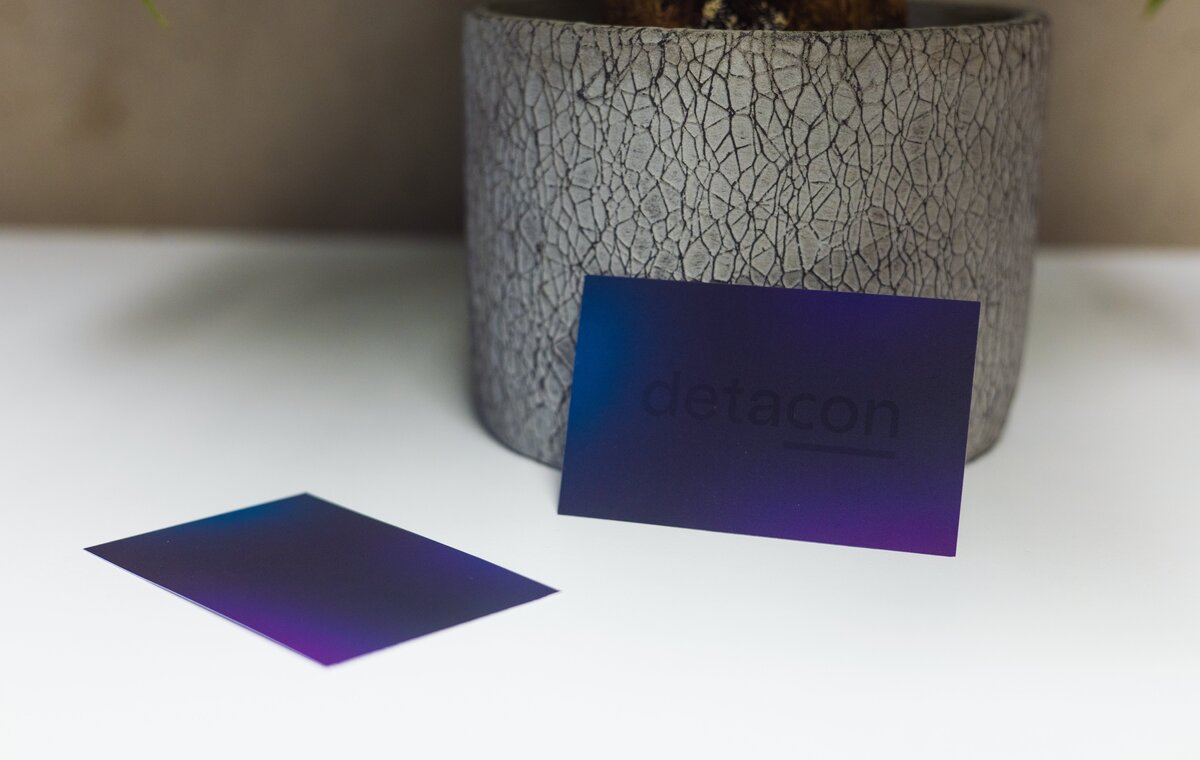 Zwei dunkelblau-violette Visitenkarten mit dem detacon-Logo liegen auf einem weißen Tisch vor einem grauen Steintopf.