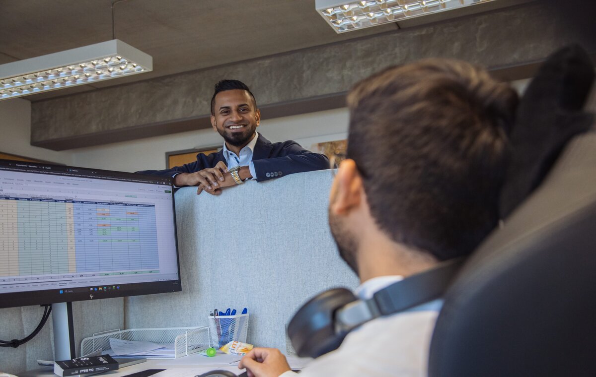 Ein Mitarbeiter lächelt über eine Trennwand hinweg zu einem Kollegen, der an einem Monitor mit Tabellenkalkulation arbeitet.