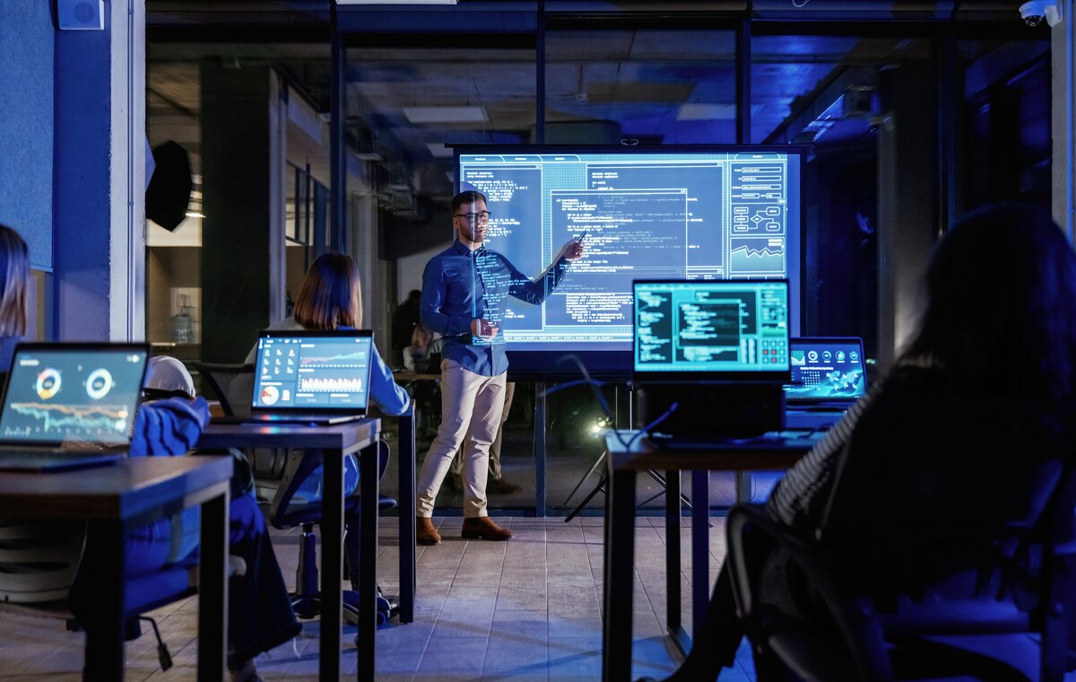 In einem abgedunkelten, modern ausgestatteten Schulungsraum präsentiert ein Referent technische Datenvisualisierungen und Programmcode auf einer großen Projektionswand. Im Vordergrund sind die Silhouetten von Kursteilnehmern an ihren Laptops zu sehen.