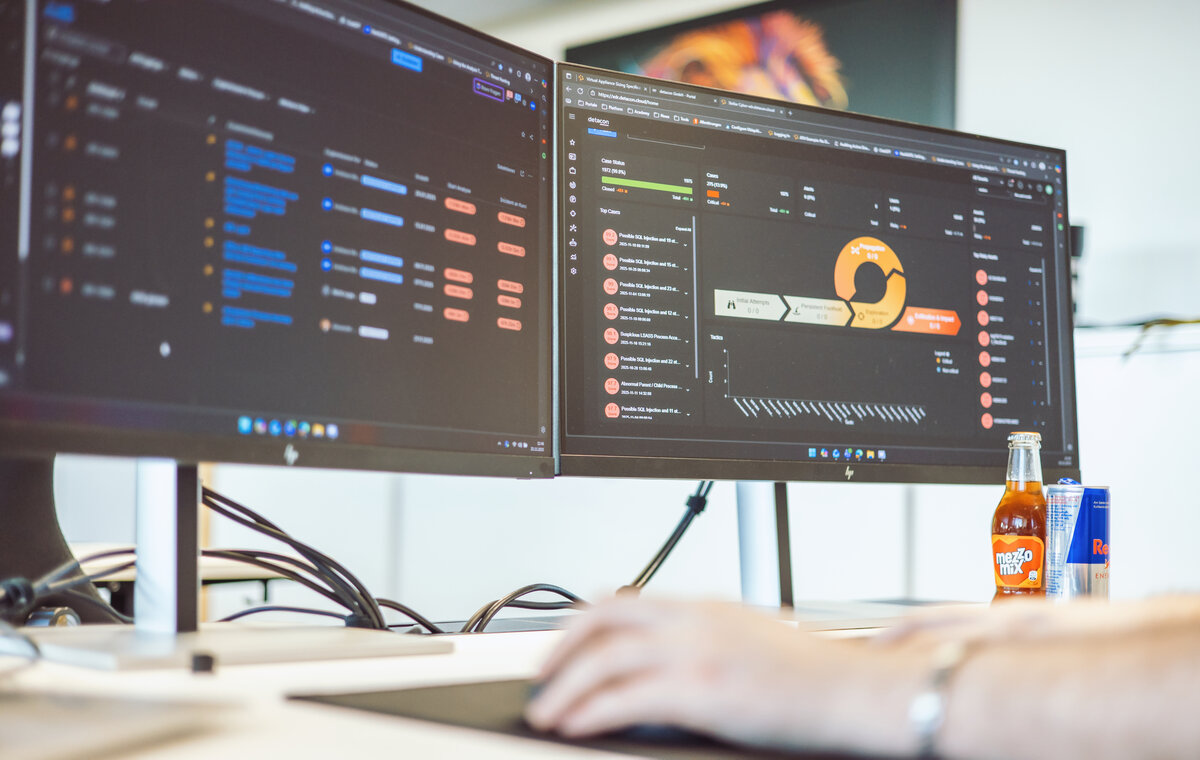 Zwei große Computerbildschirme auf einem Schreibtisch zeigen komplexe Dashboards mit Sicherheitsanalysen, Bedrohungsmetriken und Prozessdiagrammen in dunklem Design.