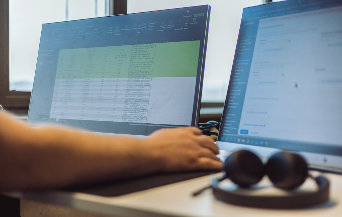 Nahaufnahme eines Arbeitsplatzes mit zwei Monitoren. Auf dem linken Bildschirm ist eine detaillierte Tabellenkalkulation mit grün markierten Zeilen zu sehen, auf dem rechten ein Web-Interface. Im Vordergrund liegt ein Headset.