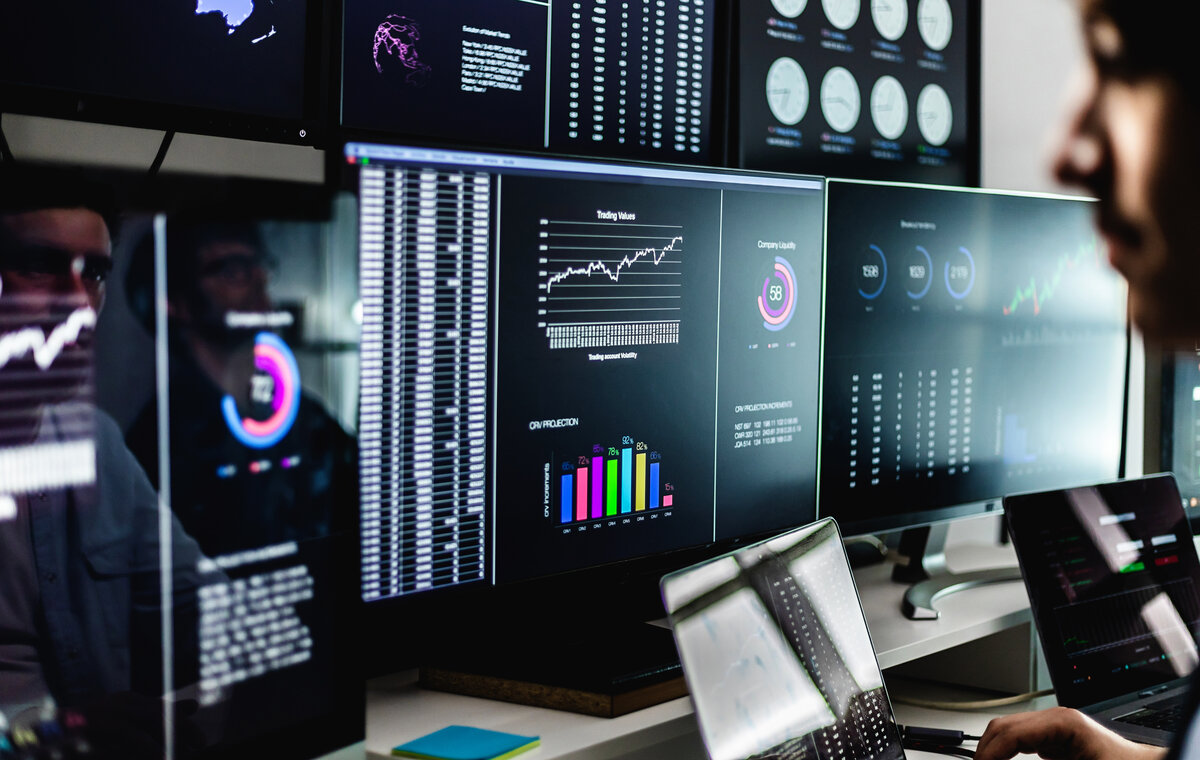 Nahaufnahme eines IT-Überwachungszentrums mit mehreren Bildschirmen, die komplexe Datenvisualisierungen, Kursdiagramme und geografische Sicherheitsmetriken in einem abgedunkelten Raum zeigen.