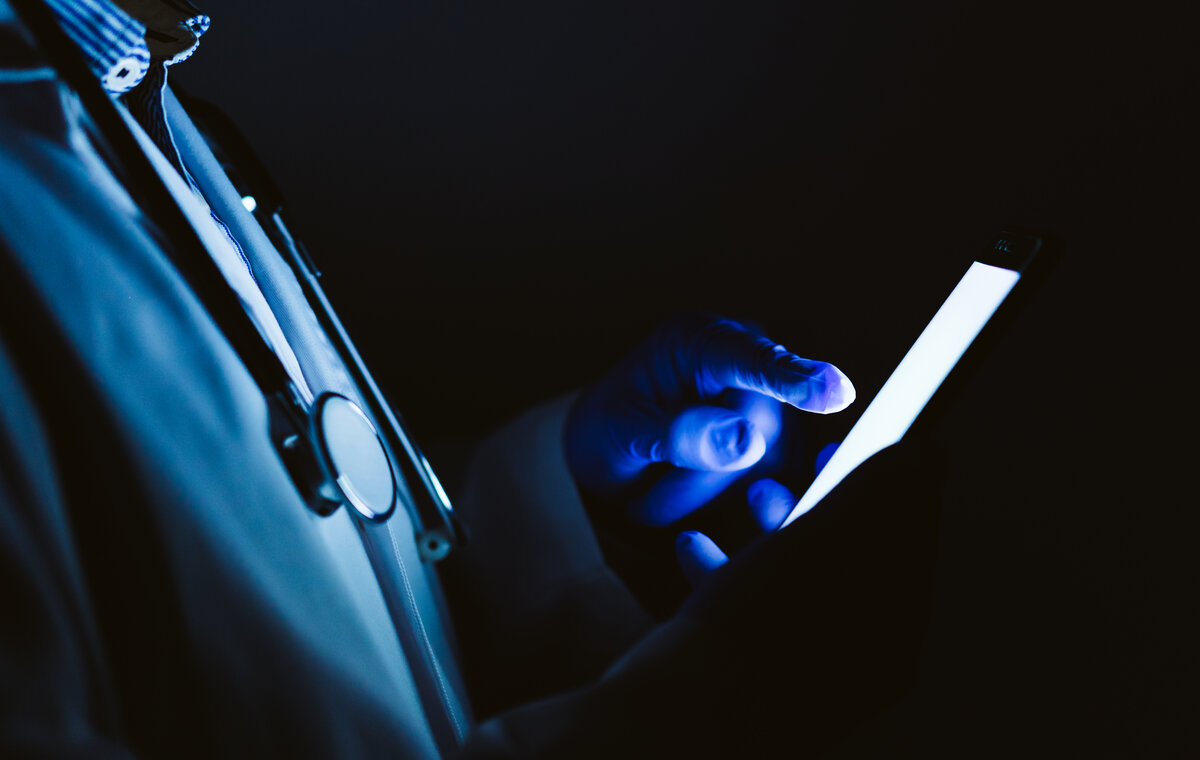 Nahaufnahme eines Mediziners im Kittel, der in dunkler Umgebung sicherheitskritische Daten auf einem leuchtenden Smartphone abruft.