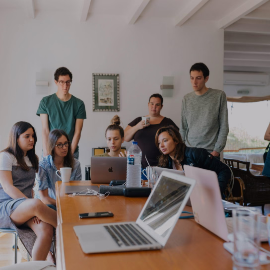 Group of seven young adults gathered around a table, attentively looking at laptop screens in a bright room.