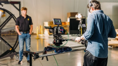 Camera operator filming with a professional camera setup on a slider, with a man in a black shirt standing in the background in a studio.
