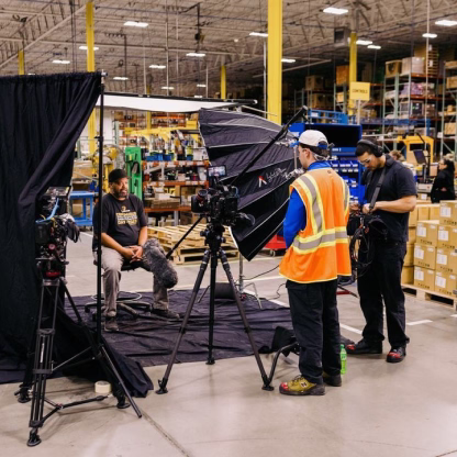Film crew in a warehouse setting recording an interview with a man seated on a stool surrounded by professional camera and lighting equipment.