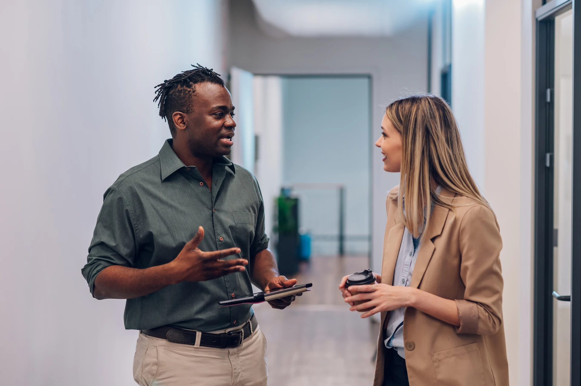 Two colleagues standing in a hallway, engaged in a conversation; one holds a tablet and the other a coffee cup.