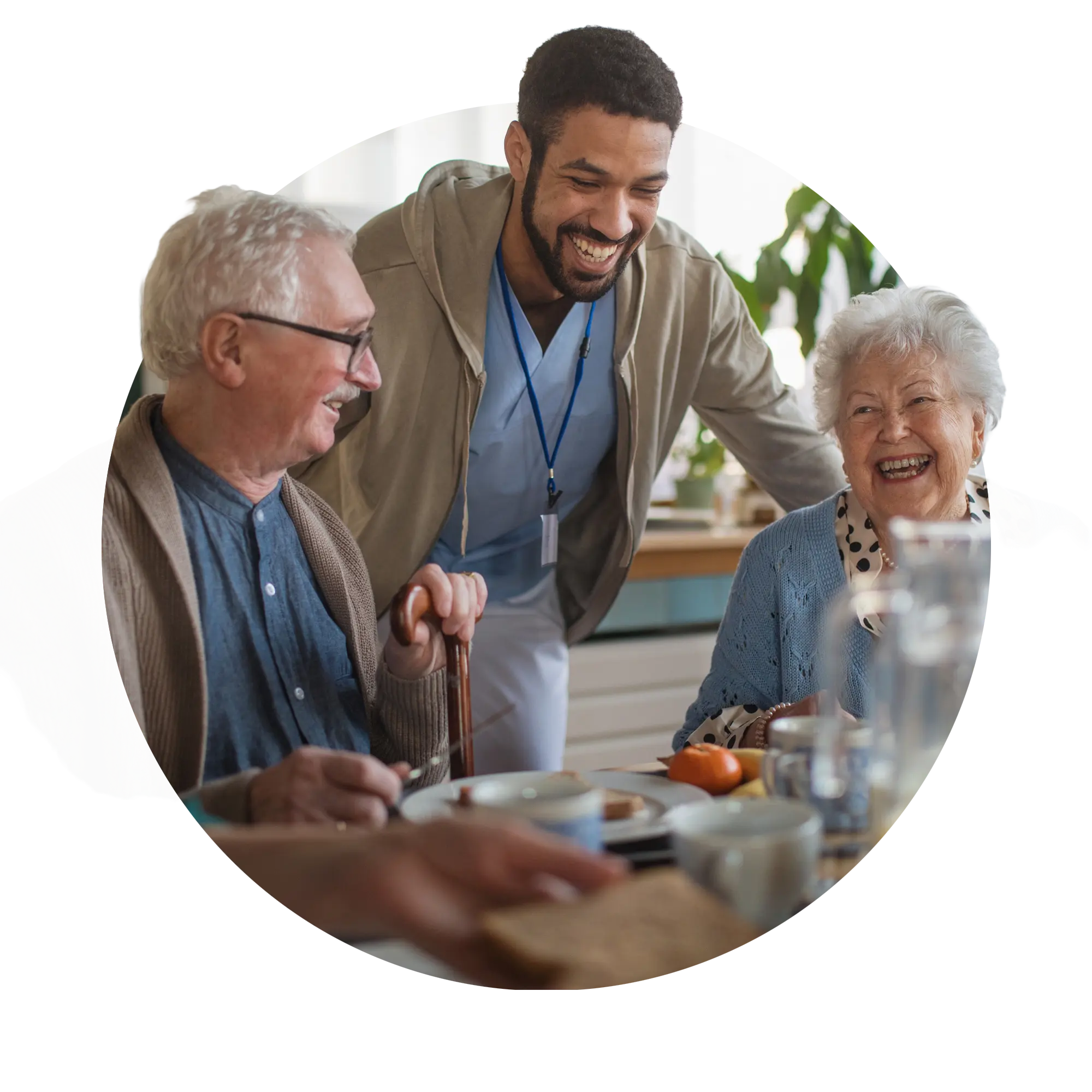Smiling caregiver talking with an elderly man and woman sharing a meal at a table.